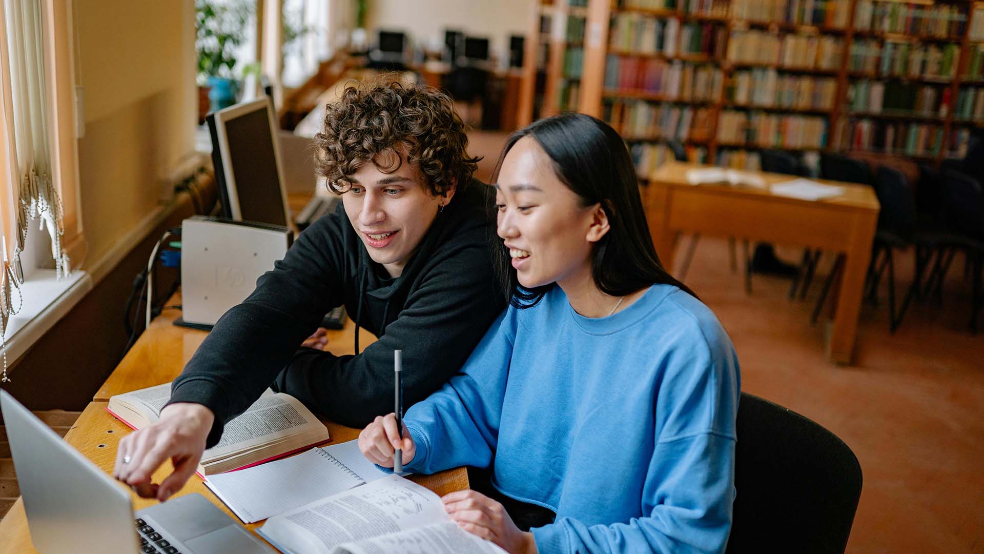 two friends studying in a library using laptop and books