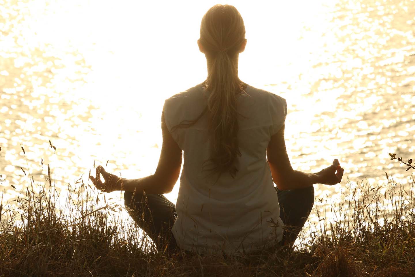 Woman sits on grass in front of a body of water at sunrise or sunset, while meditating with her legs crosses and arms balanced on her knees.