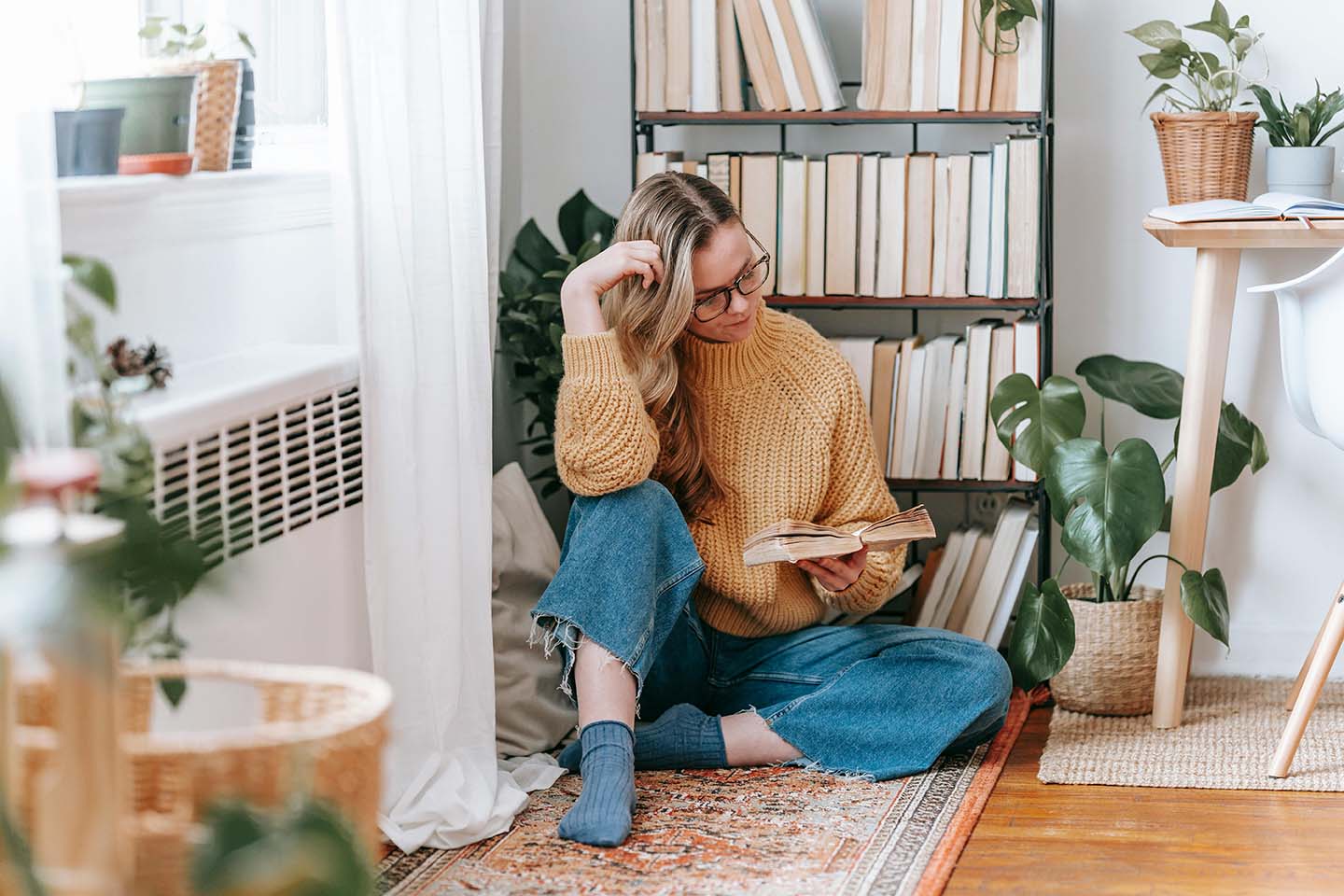 woman in jeans sitting on a wood floor reading a book in front of a bookcase